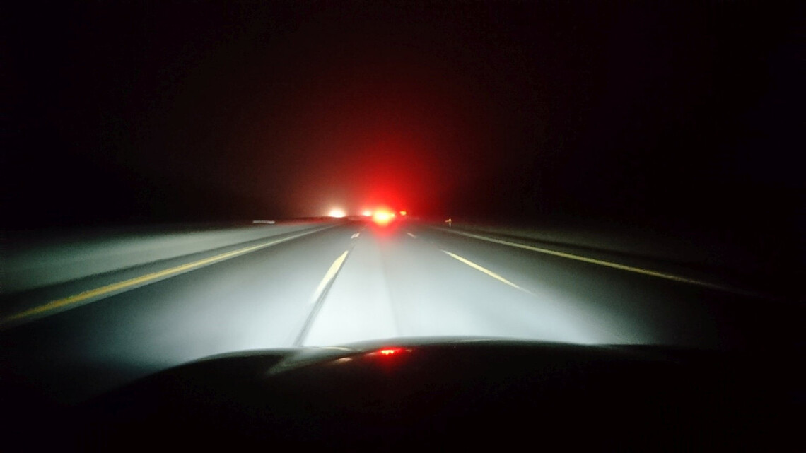 Pohľad z kapoty auta idúceho po nočnej diaľnici v ľahkej hmle. Auto pred ním v diaľke má zapnuté hmlovky.