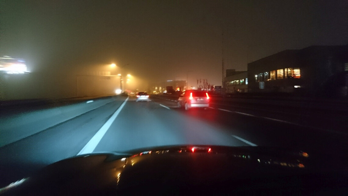 Pohľad z kapoty auta idúceho po nočnej diaľnici v ľahkej hmle. Auto pred ním má zapnuté hmlovky.